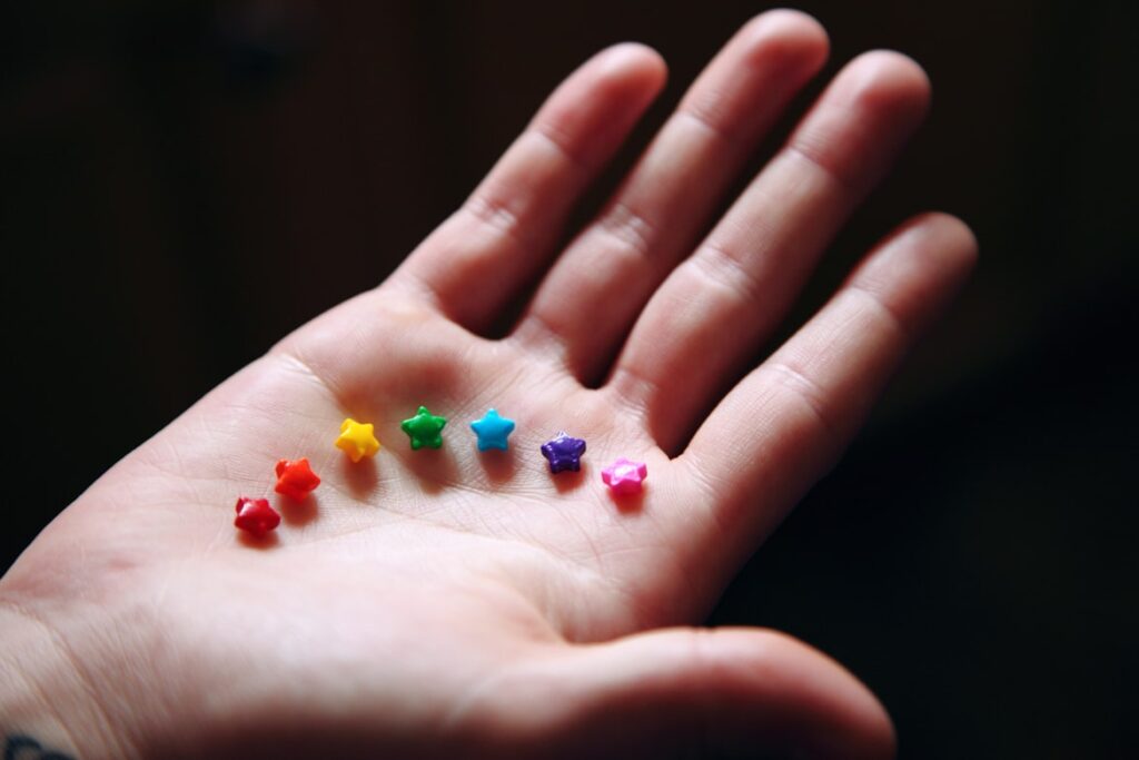 An open palm against a dark background holds seven tiny star-shaped beads arranged in a gentle arc across the skin. The stars follow a rainbow sequence from left to right: red, orange, yellow, green, blue, purple, and pink. Soft lighting highlights the glossy texture of the miniature stars.