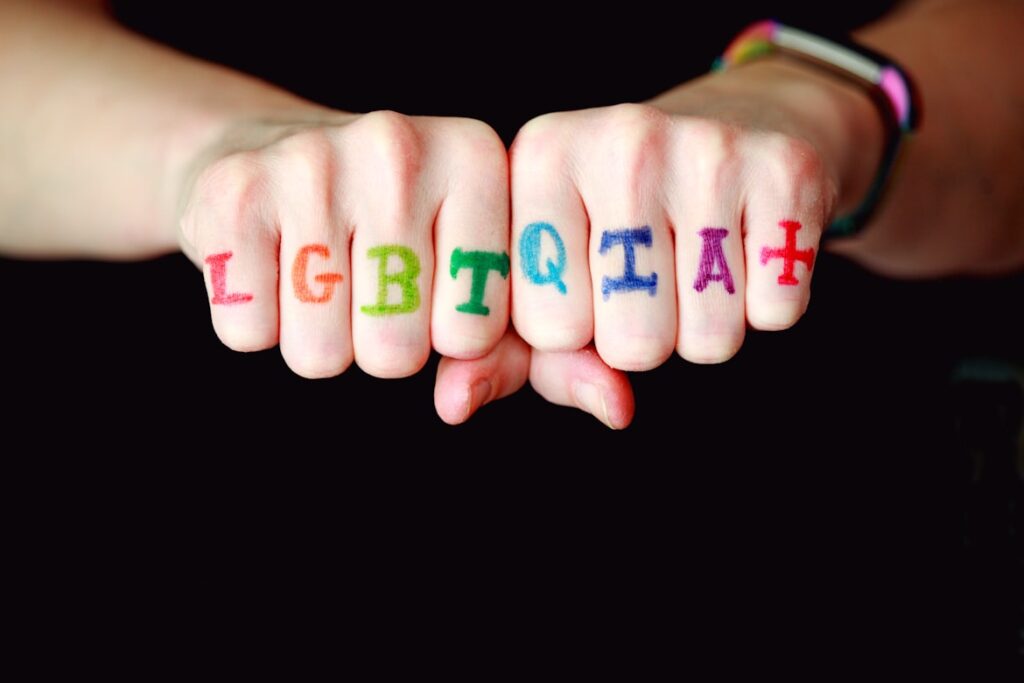 Two fists held together against a black background with the letters "LGBTQIA+" written across the knuckles in rainbow colors: L in yellow, G in green, B in teal, T in blue, Q in orange, I in light blue, A in pink, and + in red.