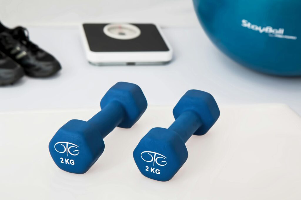 Fitness equipment arranged on a white surface, including blue 2kg dumbbells, a digital scale, an exercise ball, and black sneakers.
