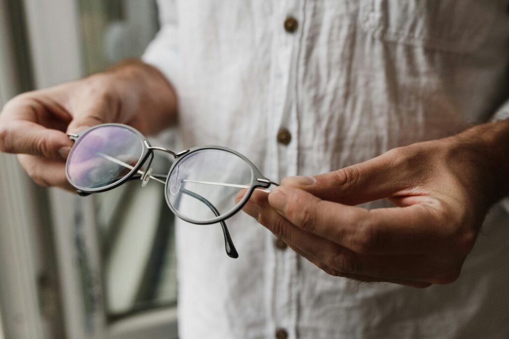 A person in a white linen shirt holding a pair of round metal-framed eyeglasses.