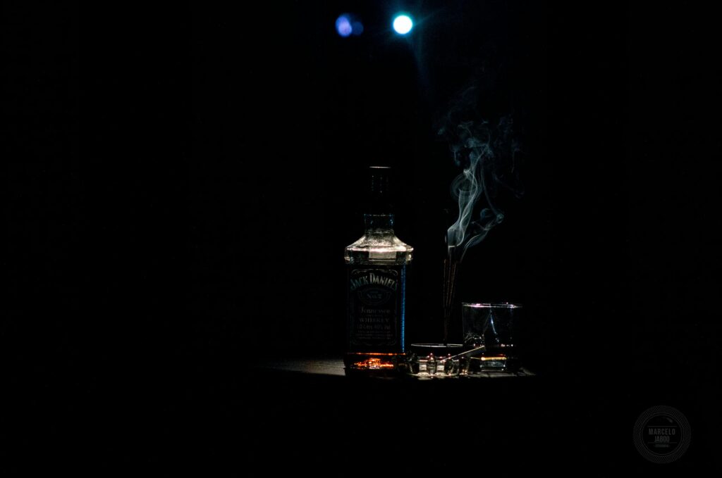 A bottle of whiskey and a lowball glass beside a smoking cigarette, dramatically lit against a dark background.