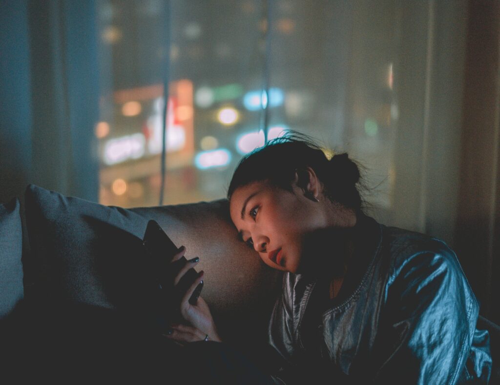 A woman lies on a couch at night, her face lit by her phone screen. Blurred city lights glow through sheer curtains behind her.