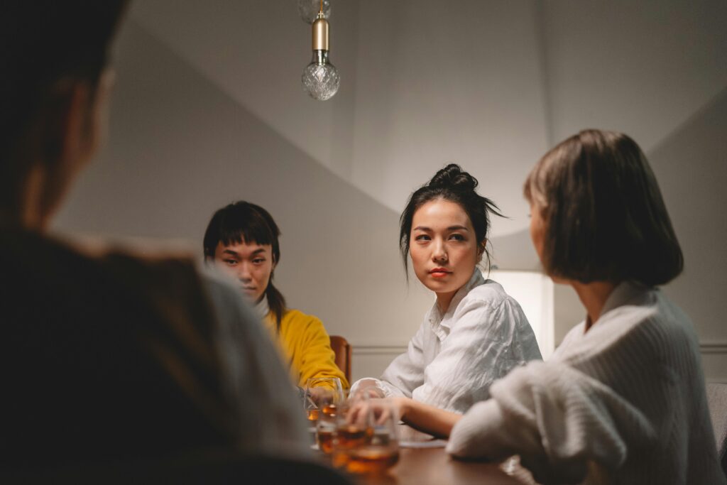 A woman with her dark hair in a bun looks directly at the camera with a serious expression while seated at a table with others. Drinks are visible on the table.
