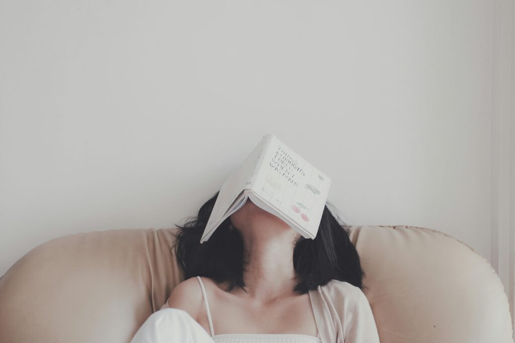 A person reclines on a cushioned seat with an open book covering their face, appearing to have fallen asleep while reading.