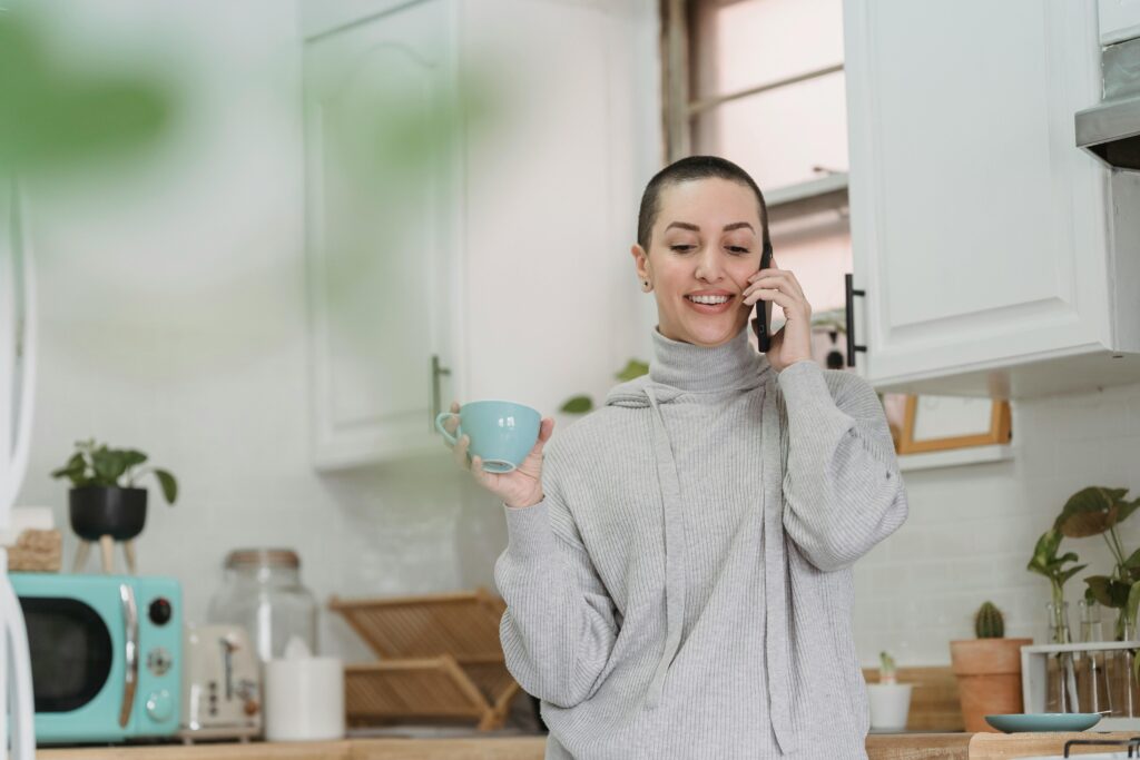 A smiling woman with short hair talks on her phone while holding a light blue mug in a bright white kitchen with plants.