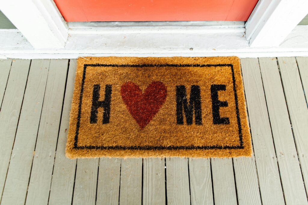 A doormat reading "HOME" with a red heart replacing the letter O, placed on gray wooden porch boards in front of an orange door.