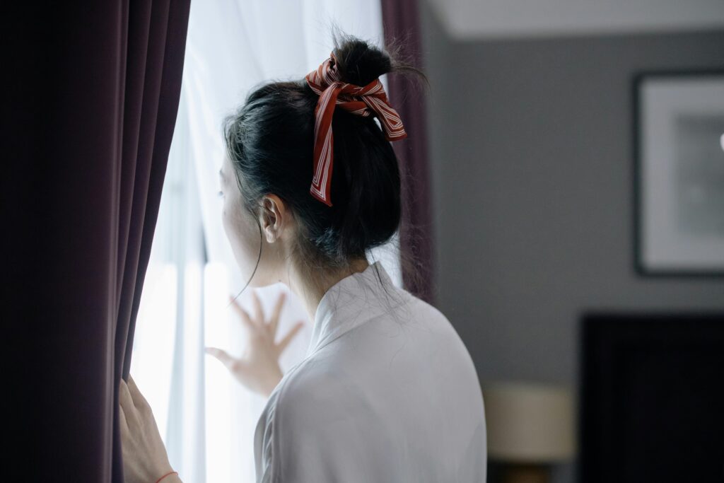 A woman seen from behind reaches toward sheer white curtains by a window. Her dark hair is in a bun tied with a striped ribbon.