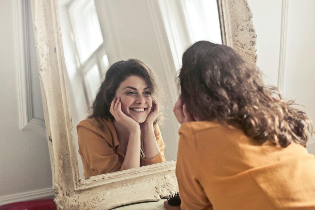 A woman with curly brown hair smiles warmly at her reflection in an ornate white-framed mirror, chin resting in her hands.
