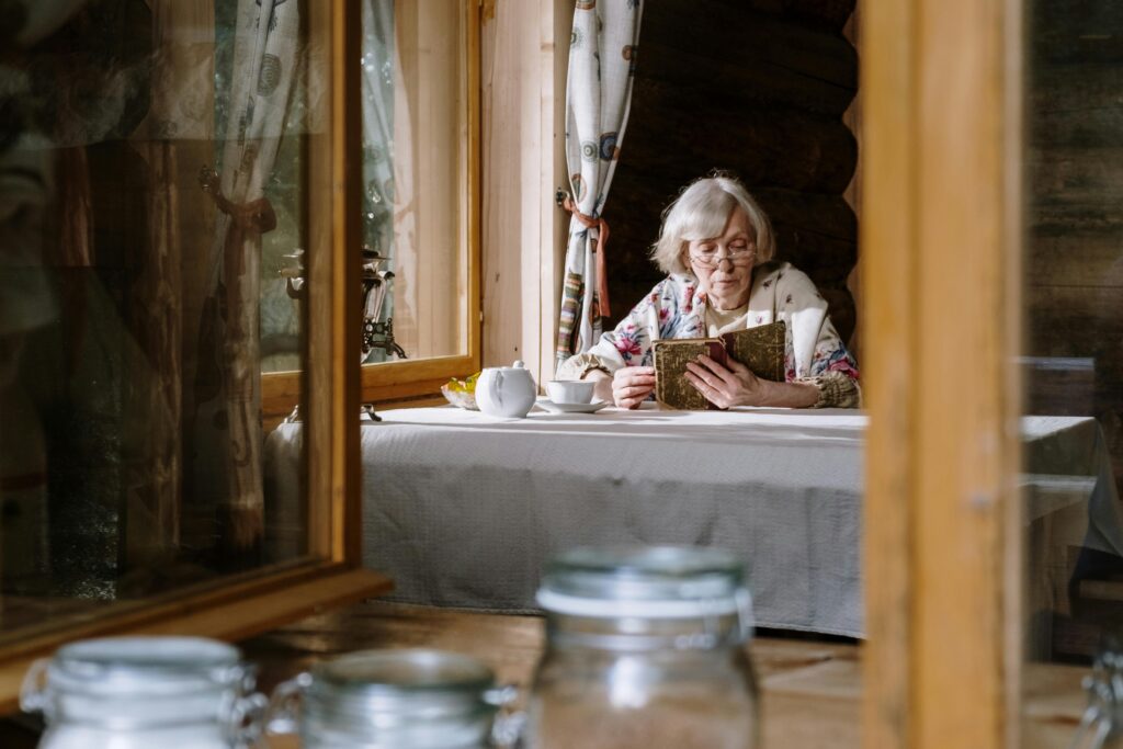 An elderly woman with white hair reads a book at a table by a window in a rustic cabin, a teapot beside her.
