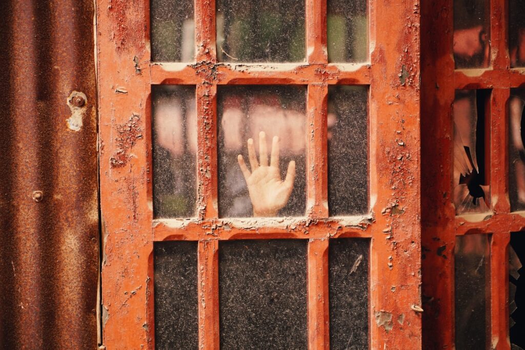 A hand presses against a grimy window behind rusted orange window pane.