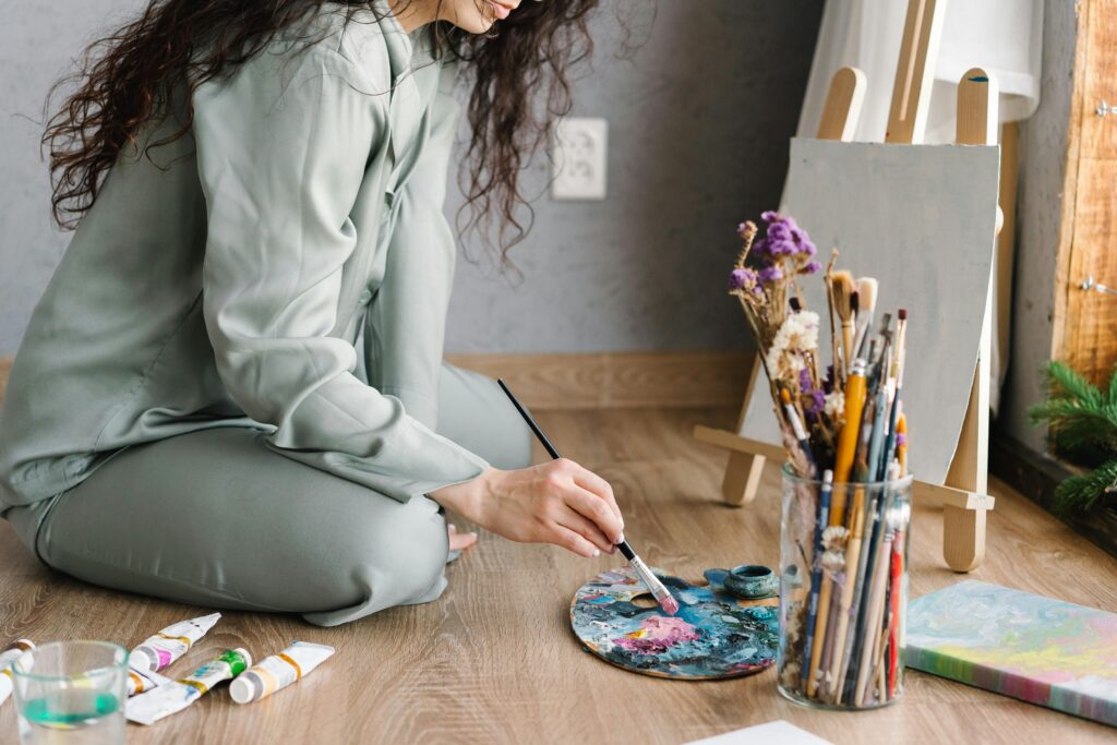 A person sits cross-legged on a wooden floor with a painter's palette, brushes, paint tubes, and a small easel nearby.