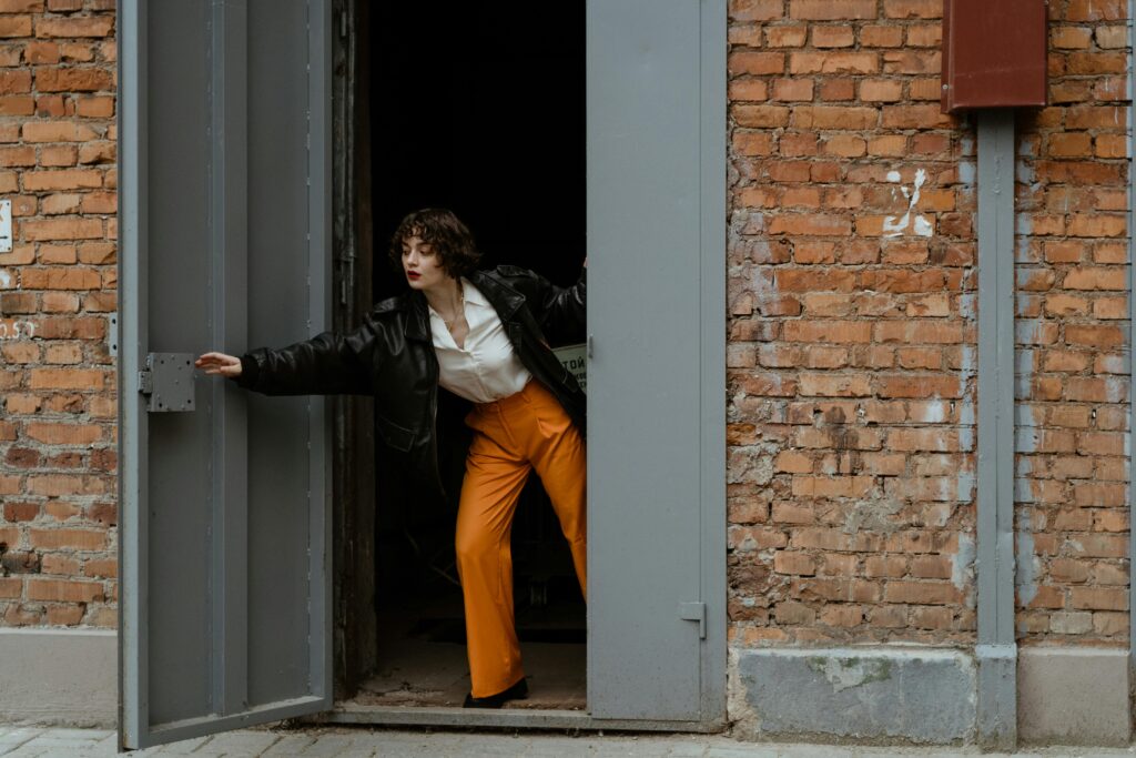 A woman in orange trousers and a black leather jacket closes a heavy door in a brick building, her curly hair catching the movement.