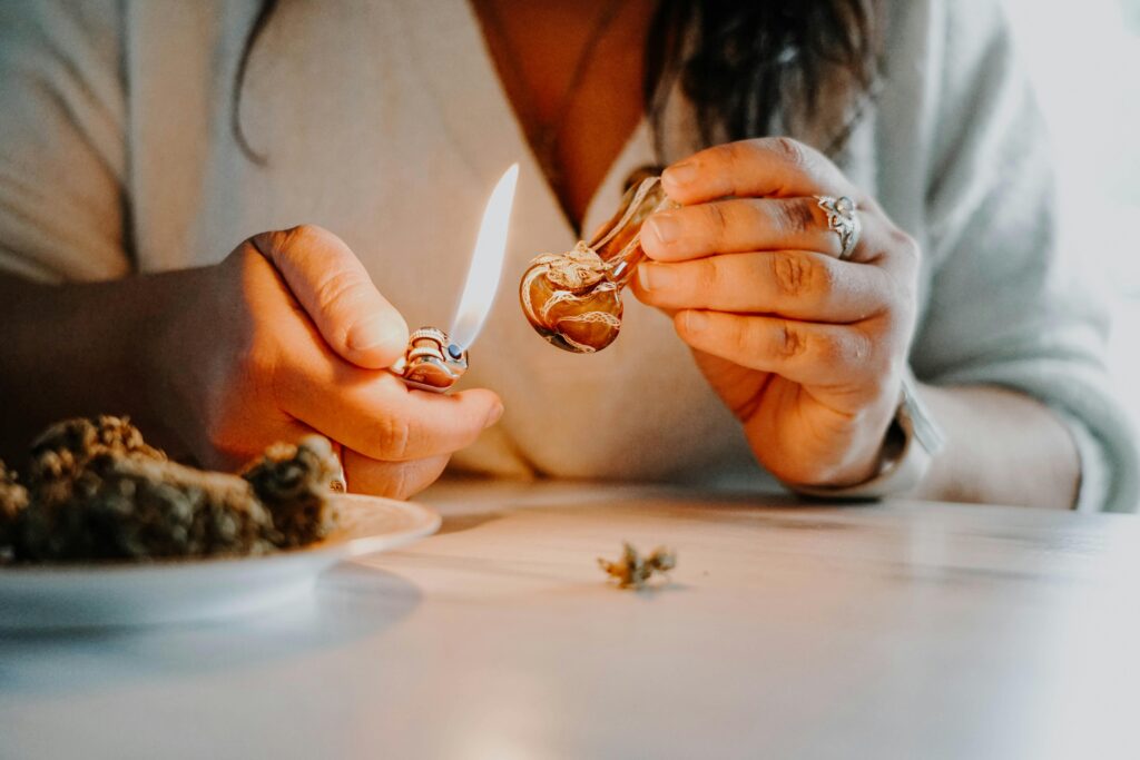 A woman in a sweater sits at a table, using a lighter to heat a small glass pipe. Cannabis buds rest on a plate beside her.