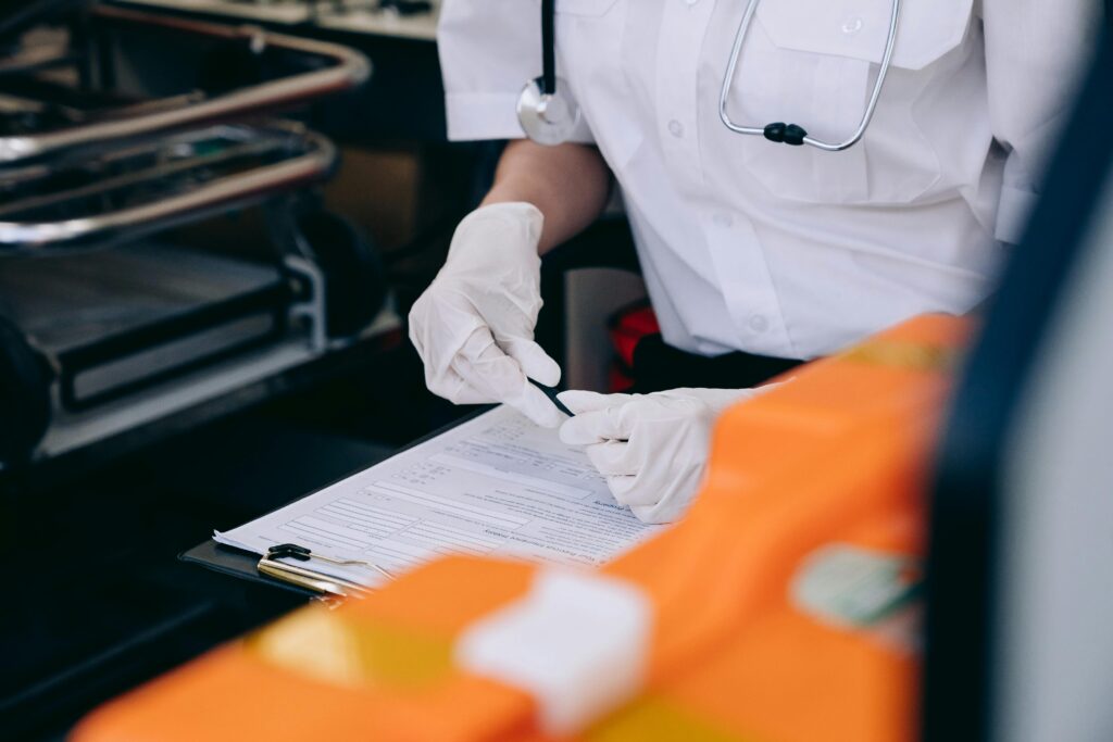 A medical professional in a white coat and gloves writes on a clipboard. A stethoscope hangs around their neck.
