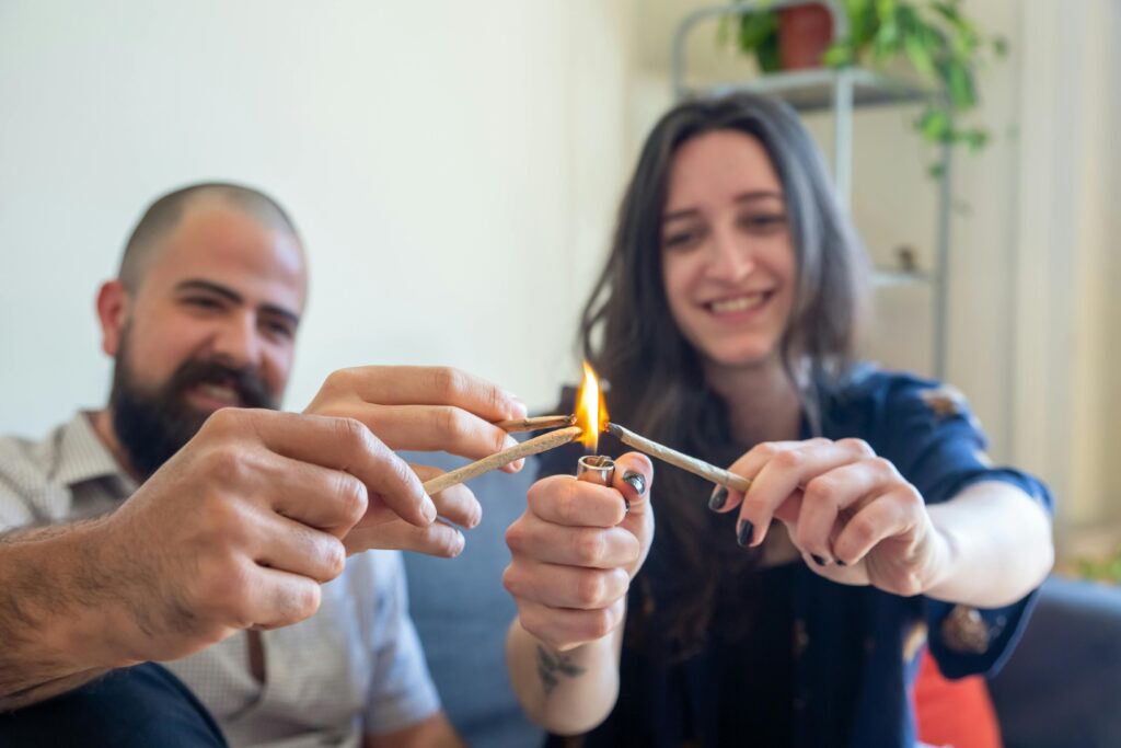 A man and woman smile as they light joints together, their hands meeting in the center of the frame.