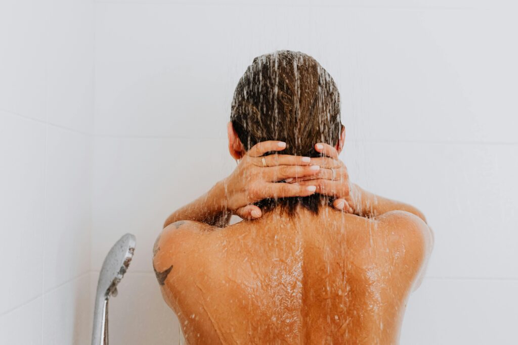 Back view of a woman showering, hands raised to their wet hair under running water.