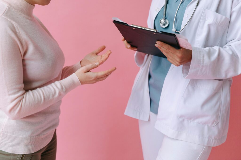 A patient in a pink sweater gestures while speaking to a doctor who holds a clipboard. Pink background.