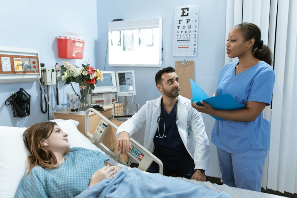 A female patient lies in a hospital bed while a male doctor and female nurse review information nearby.