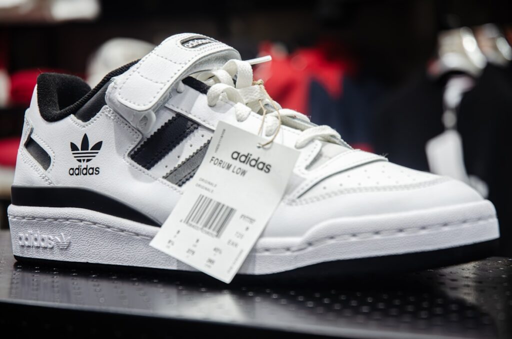 White Adidas Forum sneakers with black stripes and a price tag still attached, displayed on a store shelf.