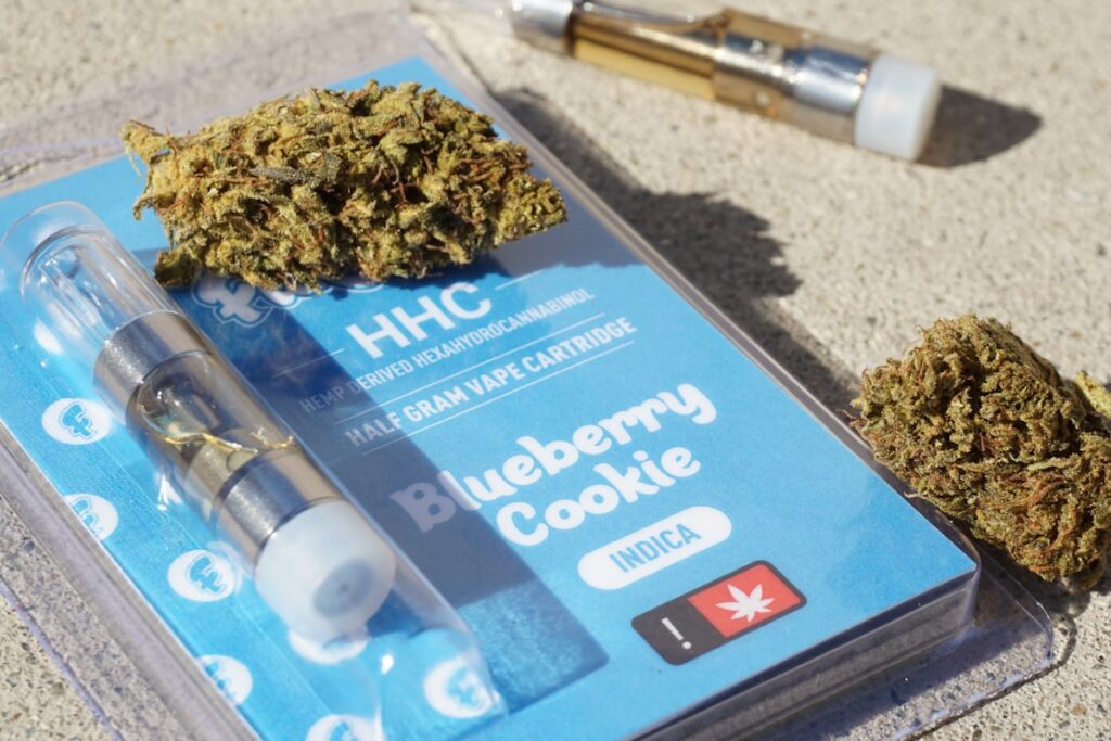An HHC vape cartridge in packaging labeled "Blueberry Cookie Indica" alongside cannabis buds and a vape pen.
