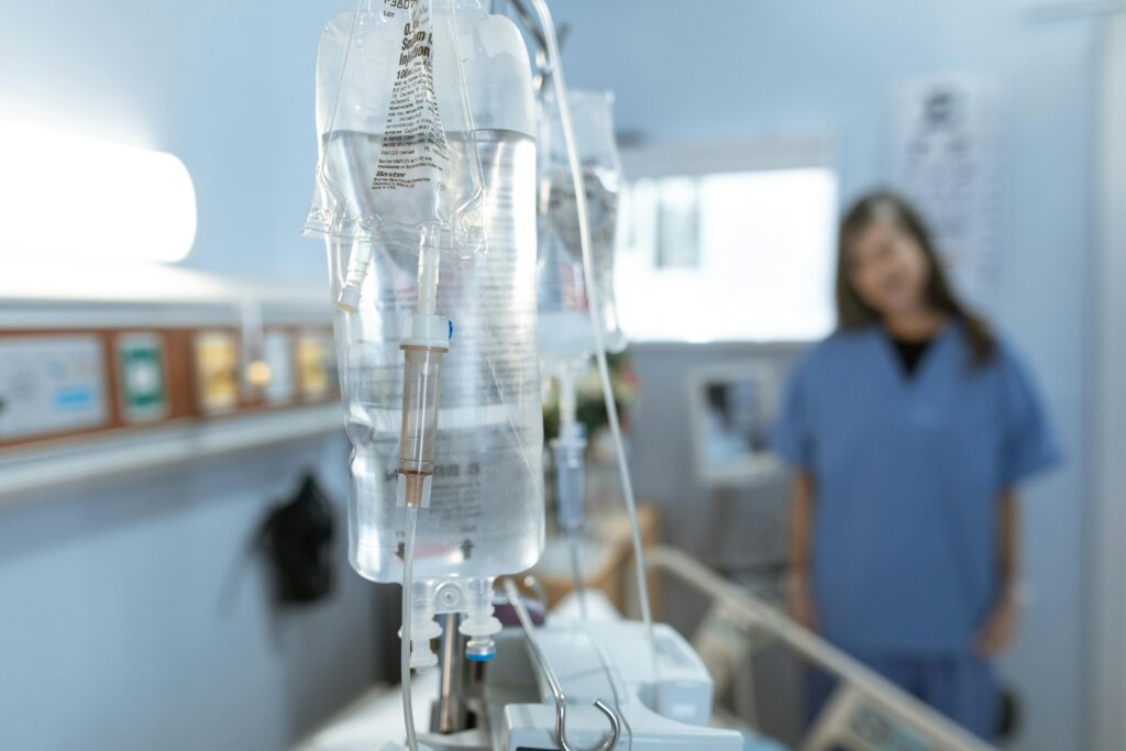 An IV drip bag hangs in focus in a hospital room, with a nurse visible but blurred in the background.