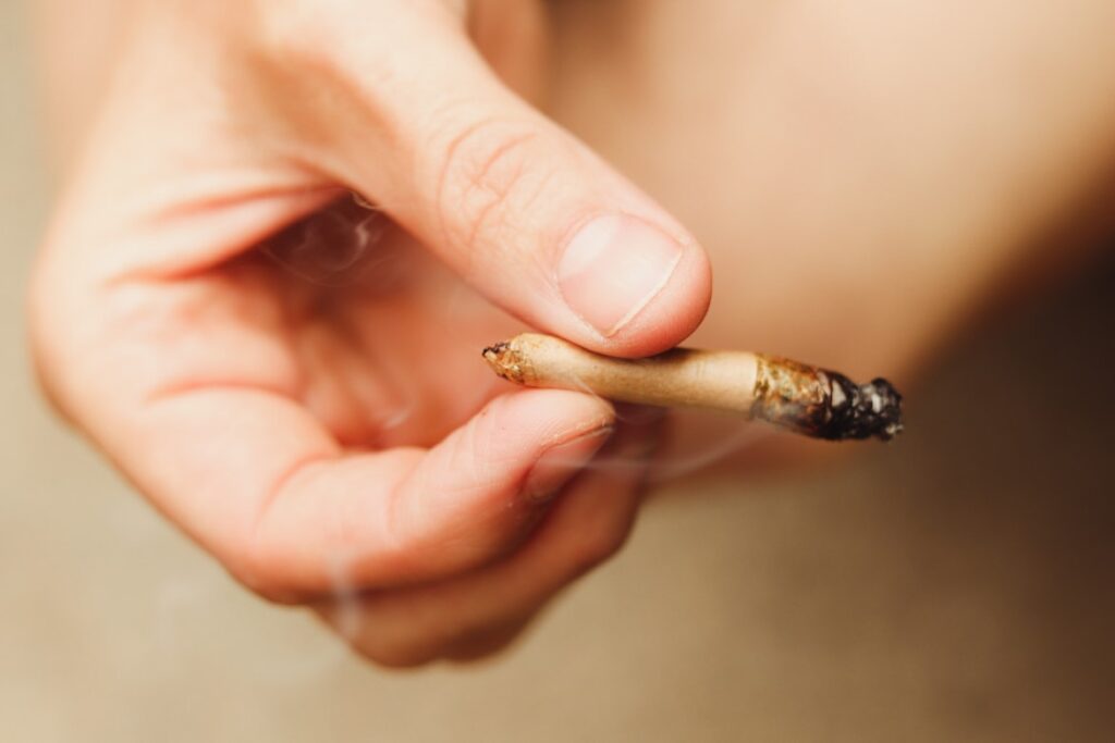 A close-up of a hand holding a lit, partially smoked joint with visible smoke.