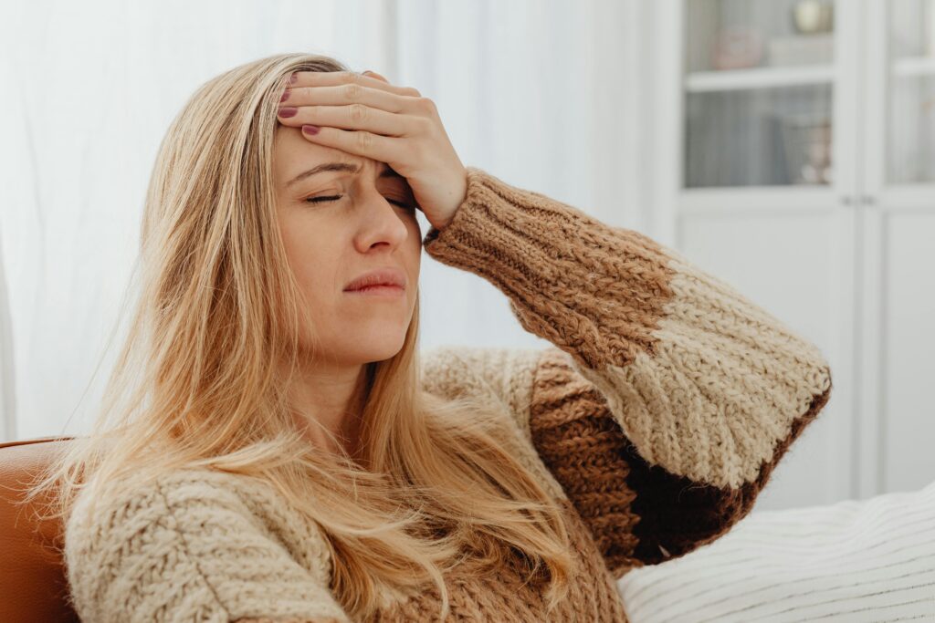 A woman with long blonde hair sits on a couch holding her forehead, eyes closed, appearing unwell.