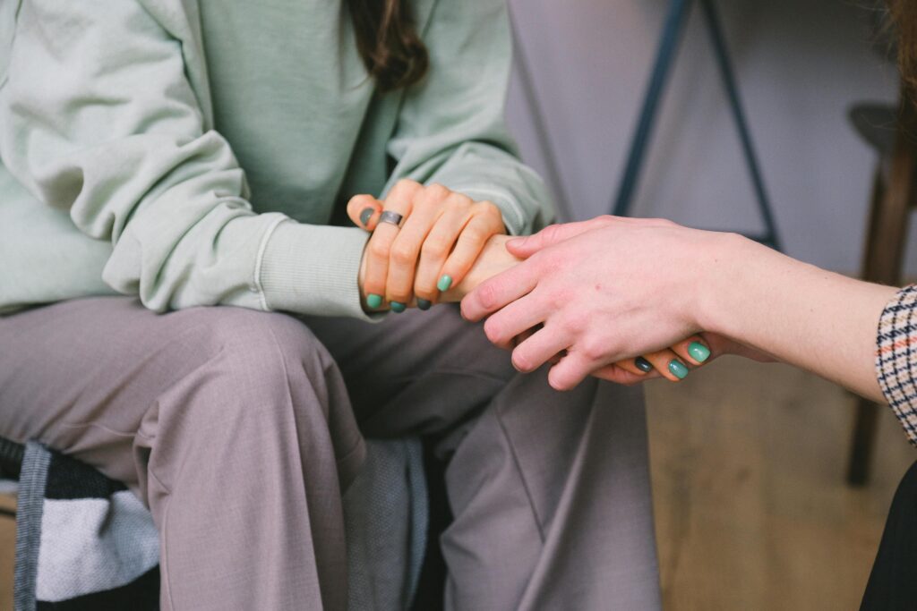 Two people sit close together, one in a sage green hoodie holding the other's hands in a gesture of comfort or support. The person offering comfort has teal nail polish.