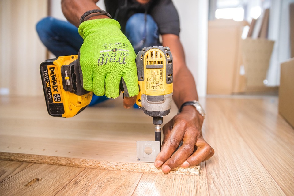 Person wearing green work gloves using a yellow DeWalt cordless drill to install wooden flooring.