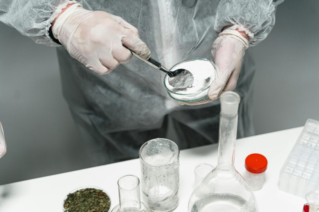 A lab technician in a white coat and gloves uses a spoon to transfer material into a dish. Beakers and containers of plant material sit on the table.