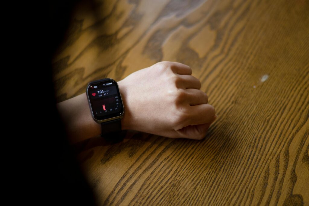 A person's hand rests on a wooden table, wearing a smartwatch with a dark band displaying health metrics on a red and black screen.