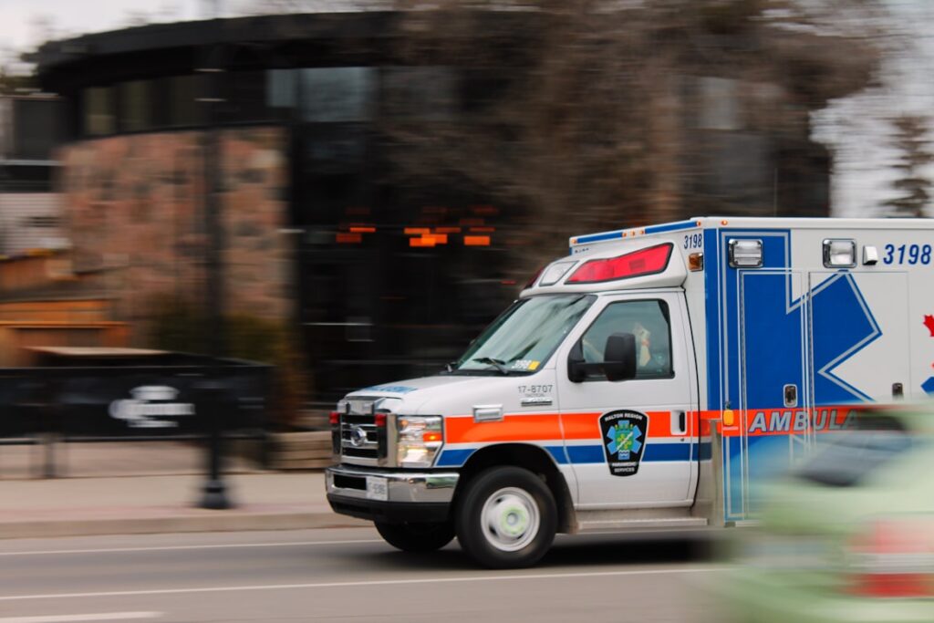 An ambulance with blue and orange markings speeds down a city street, captured with motion blur to convey urgency.