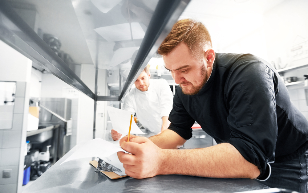 cooking and people concept - happy male chef and cook with grocery lists or bills doing inventory at restaurant kitchen