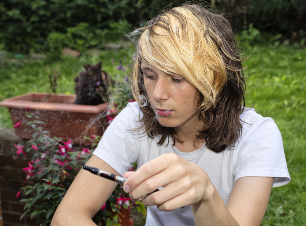 A teenage boy sitting in a garden, smoking an electronic cigarette and exhaling vapour
