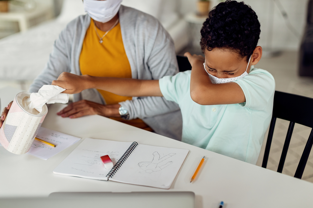 African American boy sneezing into elbow and taking a tissue from a box while learning with mother at home during coronavirus epidemic. 