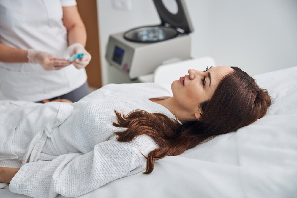 Beautiful lady in white bathrobe lying on daybed and smiling while doctor holding syringe