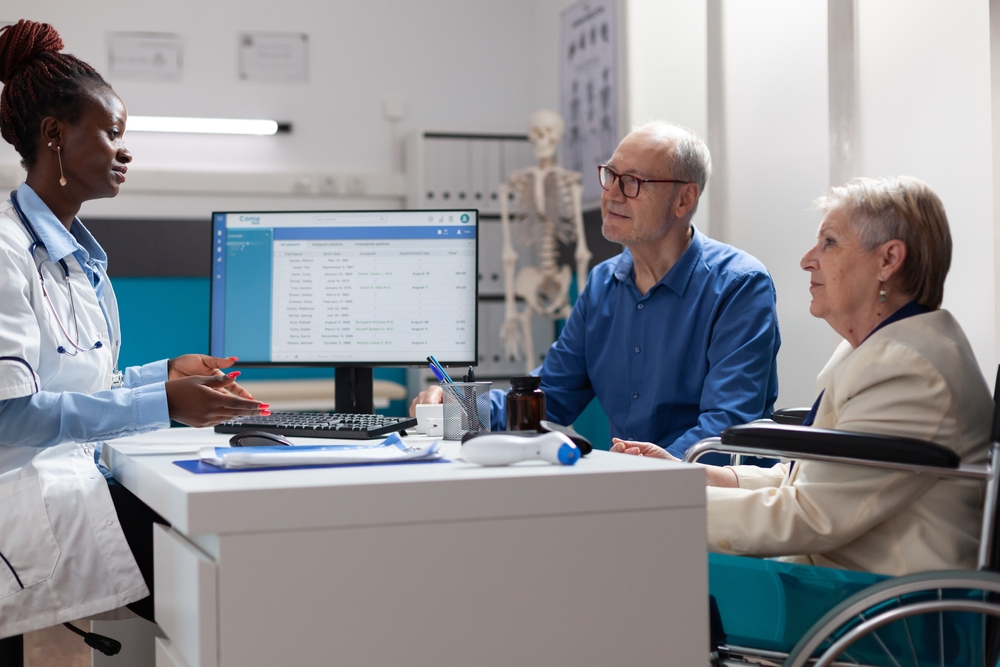 Physician doctor explaining illness expertise discussing healthcare treatment with retired senior people in hospital office. Sick old woman in wheelchair having medical appointment . Medicine concept