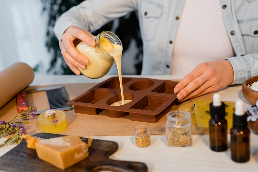 Cropped view of craftswoman pouring soap in silicone mold near flowers and essential oils on table