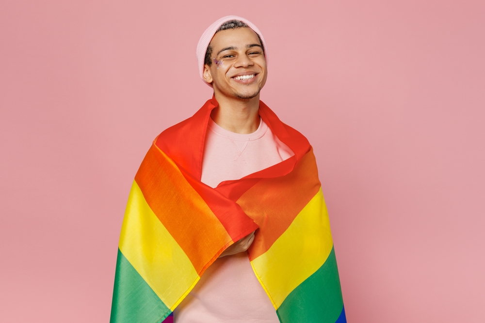 Young smiling happy gay man wear sweatshirt hat wrapped in colorful striped rainbow flag look camera isolated on plain pastel light pink color background studio portrait. Lifestyle lgbtq pride concept