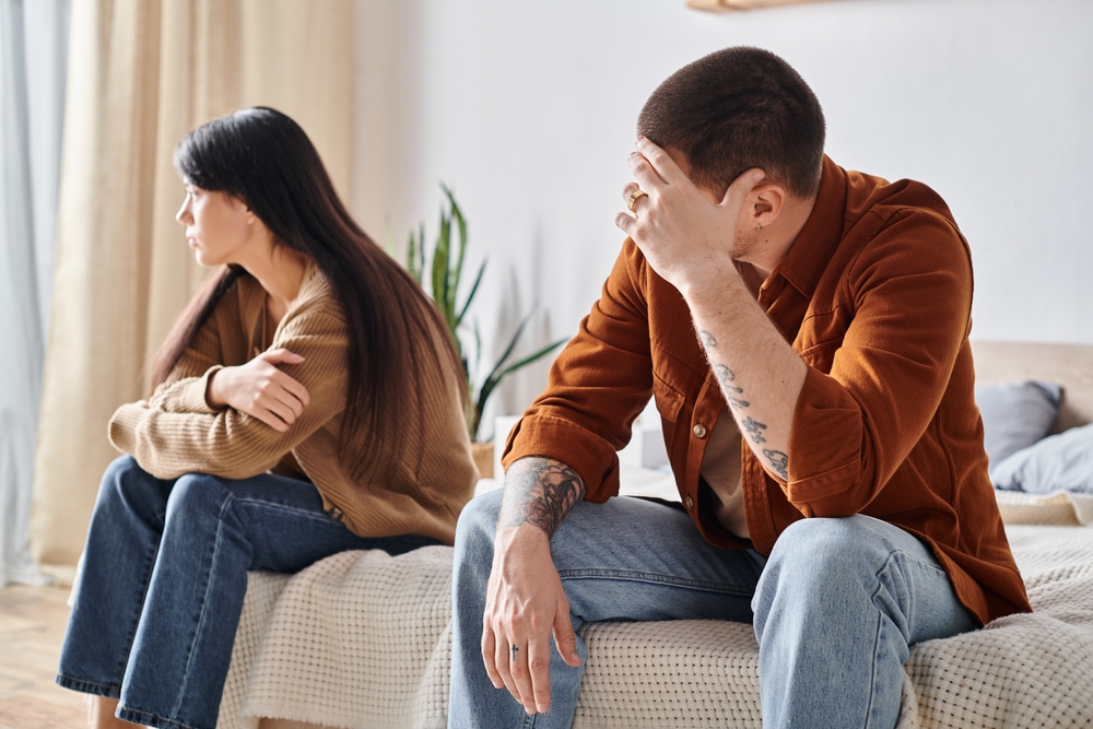 upset young interracial couple sitting in bedroom at home, relationship difficulties concept. Translation of tattoo: be faithful to the one who is faithful to you