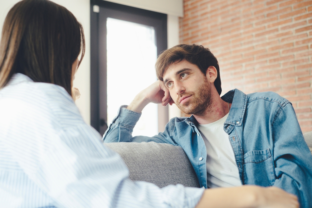 A young 25-year-old couple sits on their cozy sofa in a living room, engaged in a quiet conversation and sharing carefree moments together. Leisure and relaxation concept