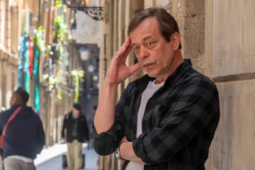 An elderly man holds his head with his hand on a busy city street.  Severe headache or lost in thought.