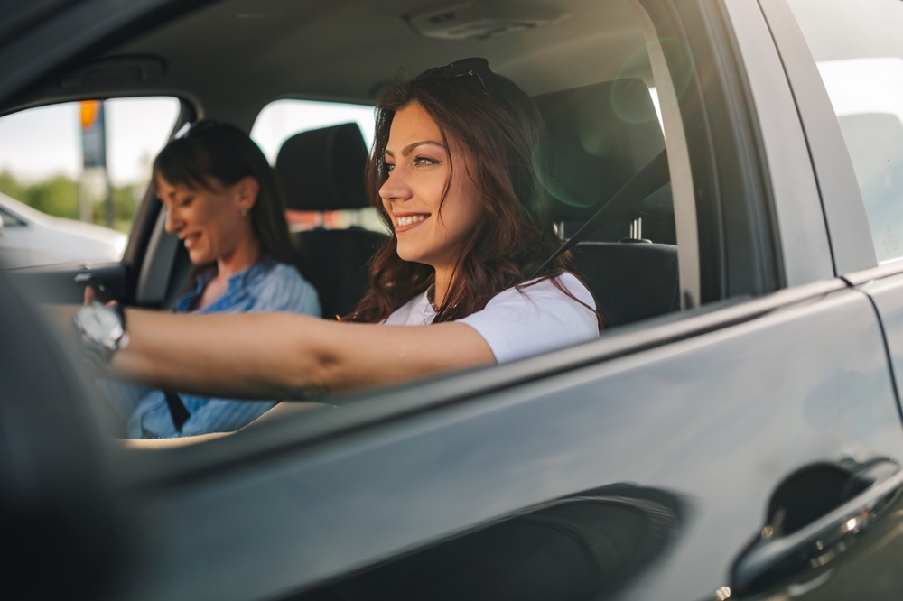 Portrait of smiling young trendy female driver sitting in a car with hands on steering wheel and driving it while her friend is sitting next to her and hanging on her phone. Fun women having road trip