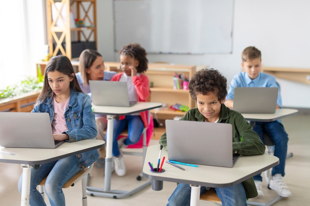 Diverse school kids having lesson, sitting at desks with laptops, studying using digital gadgets. Modern tech elementary education concept