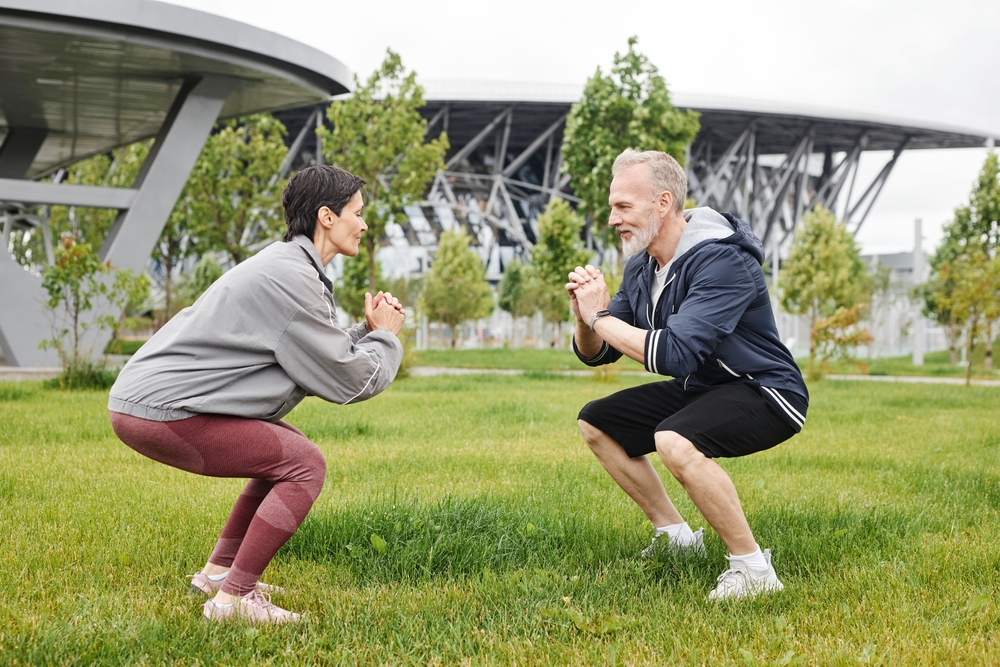 Side view of fit senior man and woman in static squat hold position working out together on green lawn at city park