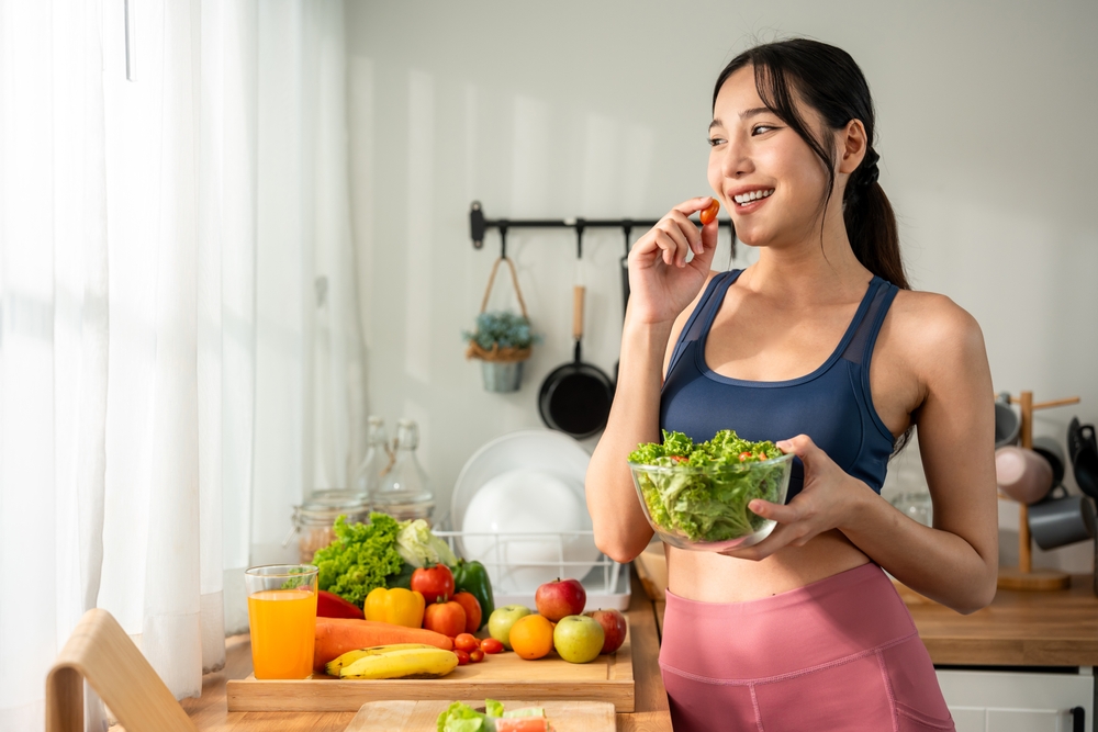 Asian sportswoman eating a bowl of salad vegetables after exercise. Beautiful young girl in sportswear enjoy eat clean healthy foods after workout for health in house. Diet and Healthy food concept.