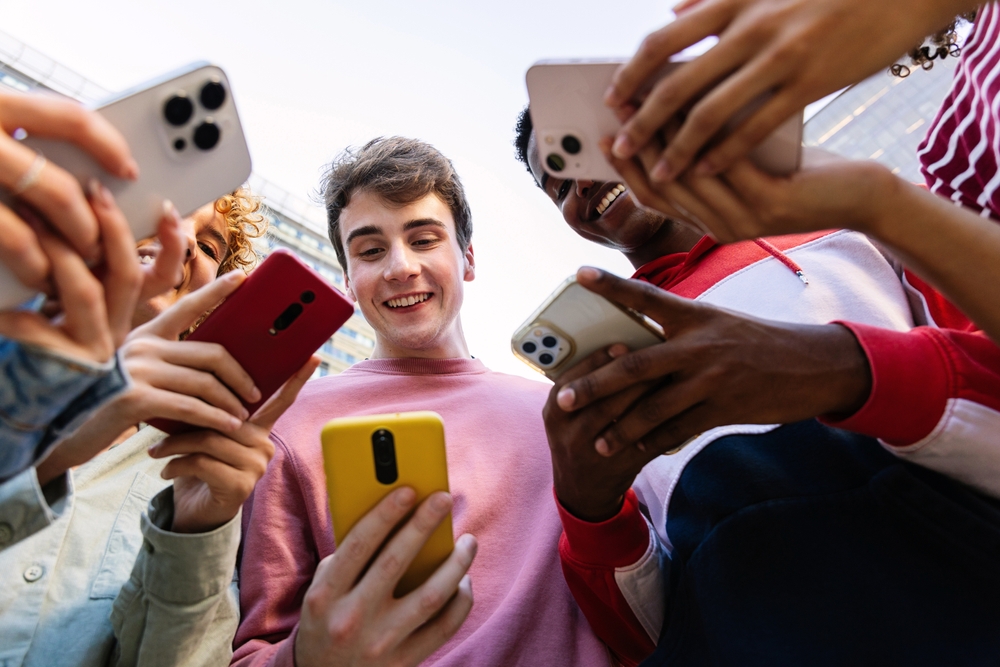 Młoda grupa nastolatków korzysta z telefonów komórkowych, stojąc w kręgu na zewnątrz. Studenci pokolenia Z uzależnieni od aplikacji społecznościowych, zakładów bukmacherskich lub gier wideo na platformie online.