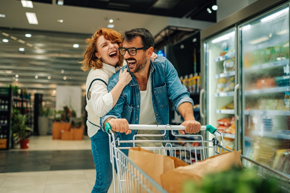 Cheerful couple is having fun in supermarket while man is pushing shopping cart and woman is embracing him, both laughing out loud