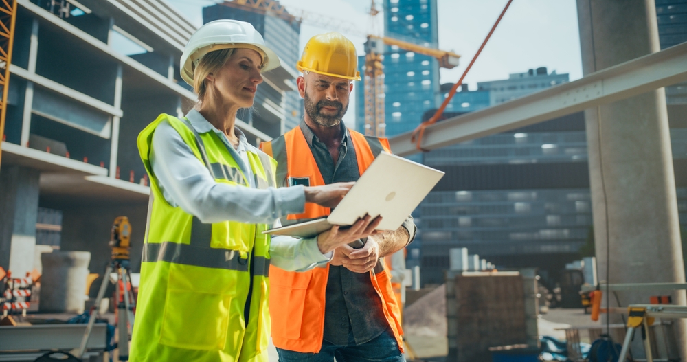 Female Civil Engineer and a Technical Inspector Having a Conversation During an Outdoors Meeting at a Building Site Area. Specialists Using a Laptop Computer to Discuss Details on a Construction Plan