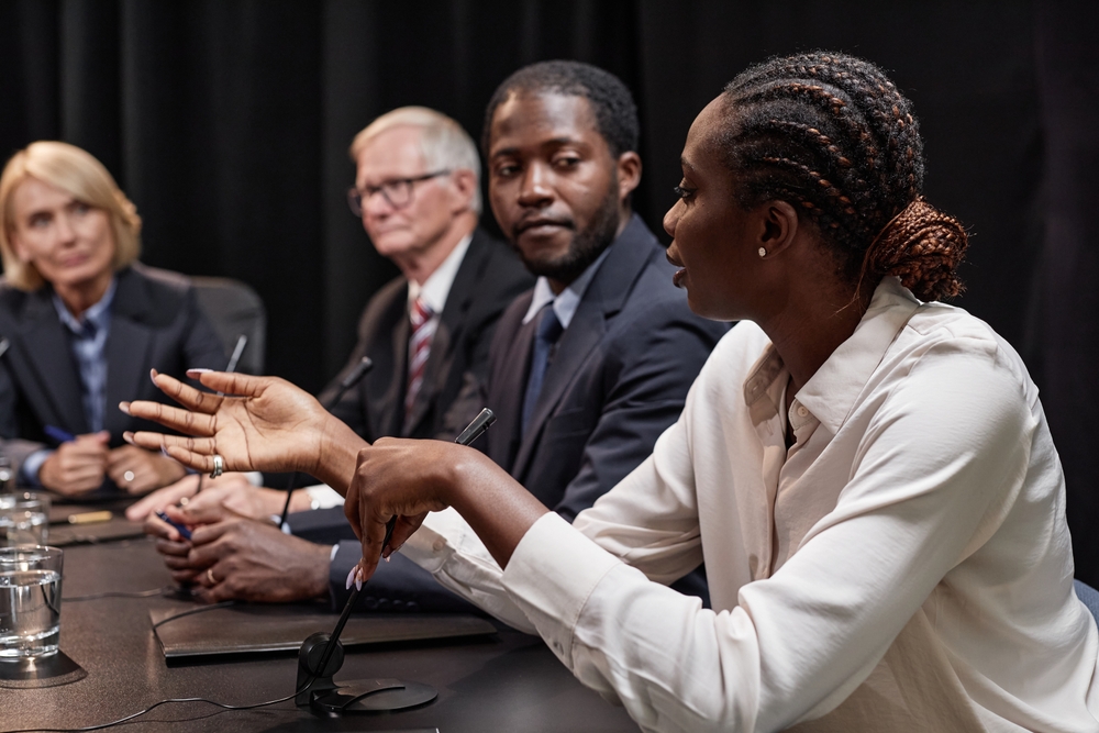 Side view of African American female politician asking colleague question speaking into microphone during committee meeting while sitting in line with other officials in conference room, copy space